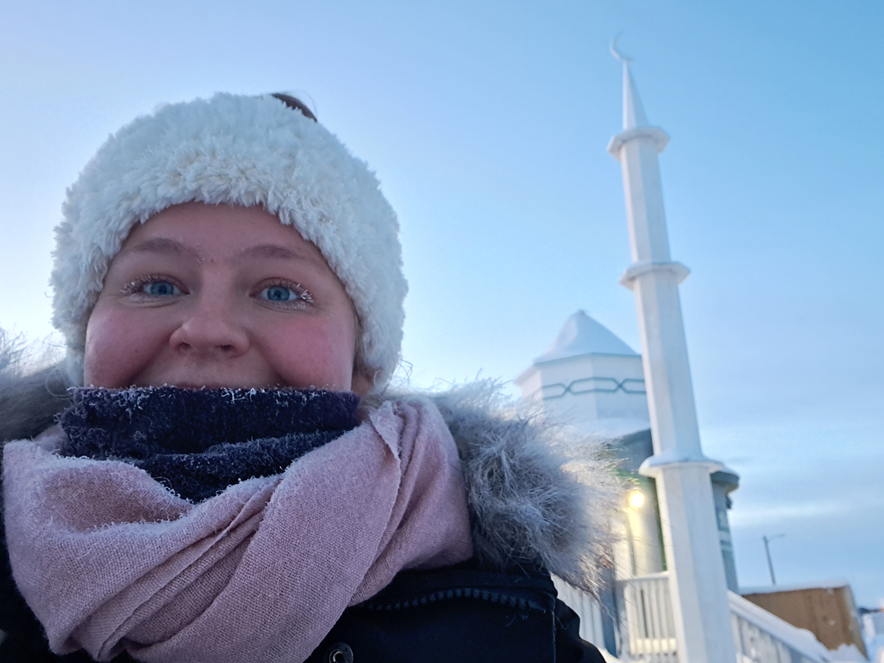 Young white woman wearing a pink scarf, white witner headband, standing in front of a white Mosque in the North